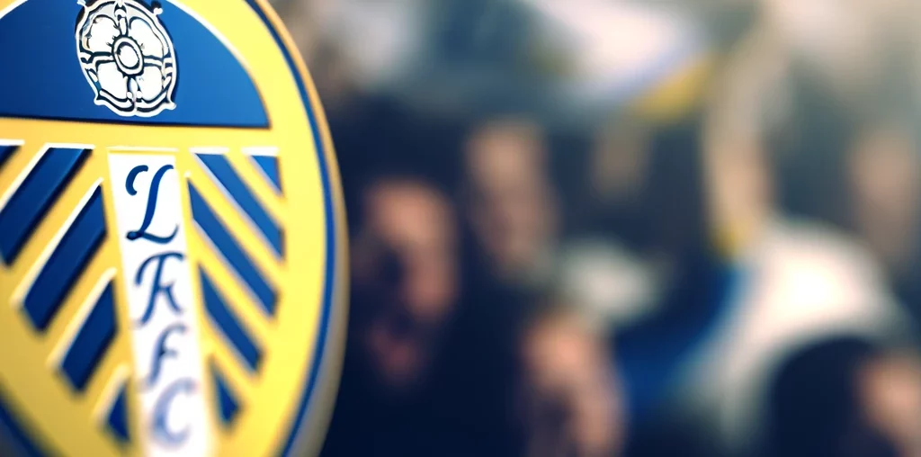 A close-up image of the Leeds United emblem. The emblem is intricately detailed, prominently featuring the team's colors of blue, yellow, and white. T
