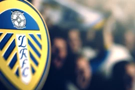 A close-up image of the Leeds United emblem. The emblem is intricately detailed, prominently featuring the team's colors of blue, yellow, and white. T
